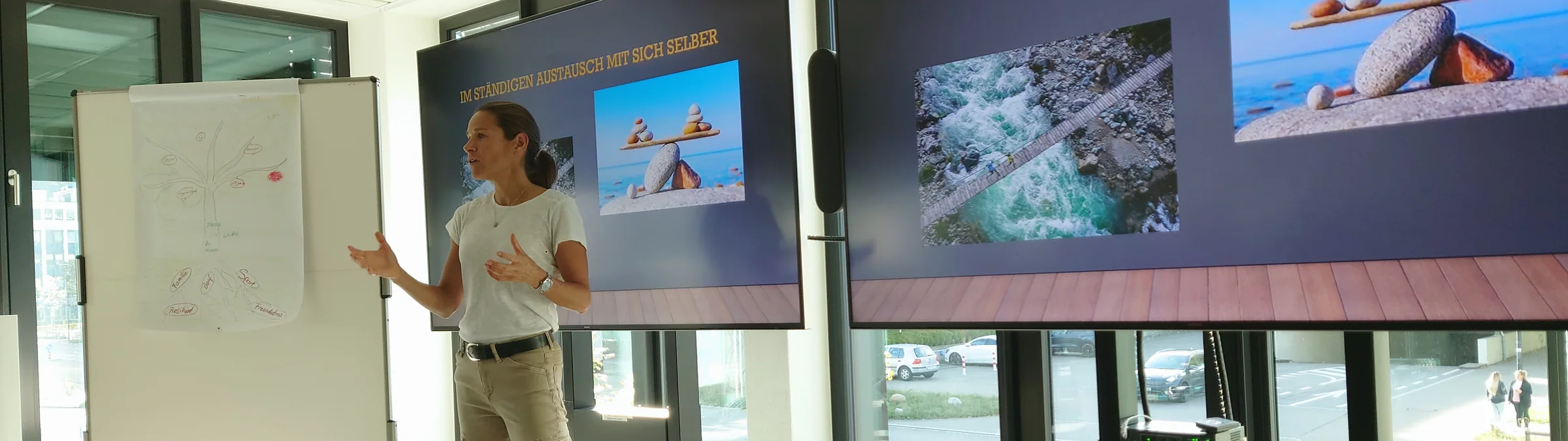 Jasmin Nunige speaking beside a flip chart, two screens with her slides behind her.