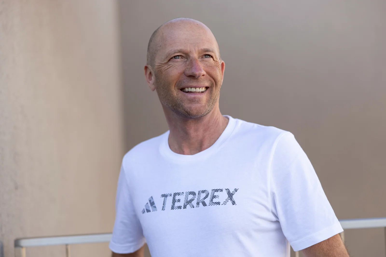 Guy Nunige in a white t-shirt laughing in front of a light wall with a railing.