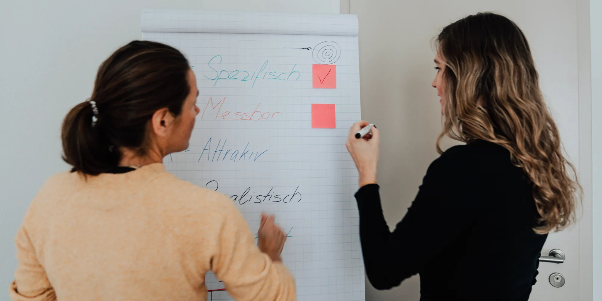 Zwei Frauen von hinten an einem Flipchart mit Zielkriterien, eine schreibt mit einem Marker neben zwei roten Haftnotizen.
