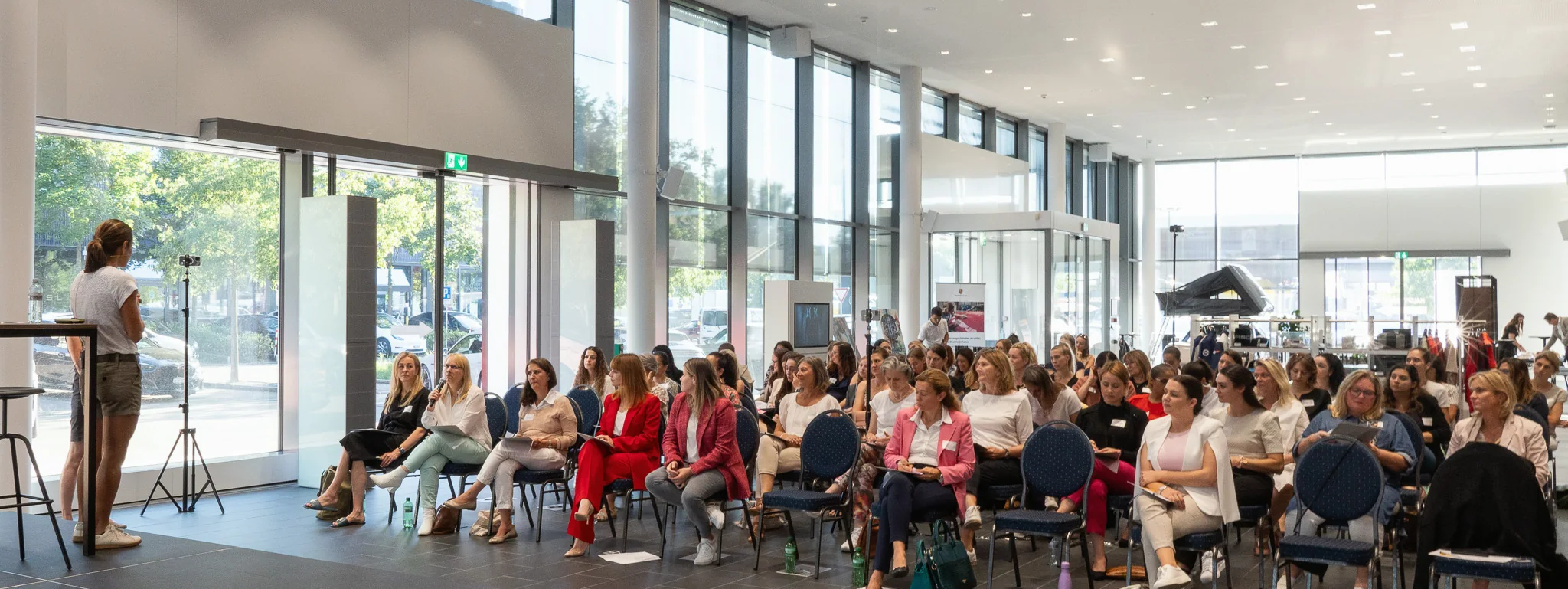 Ein voller Saal mit Publikum auf Stühlen, vorne spricht eine Person an der Glasfront.