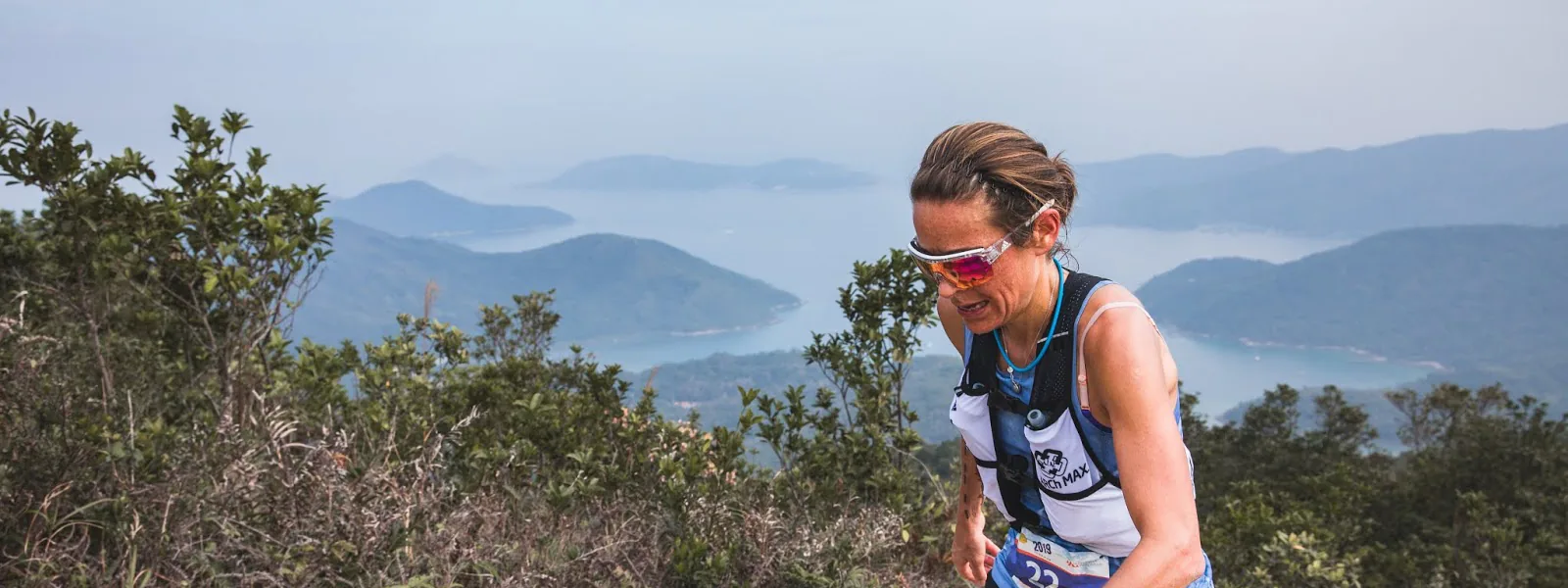 A trail runner with a race number climbing a ridge above hazy bays and hills.