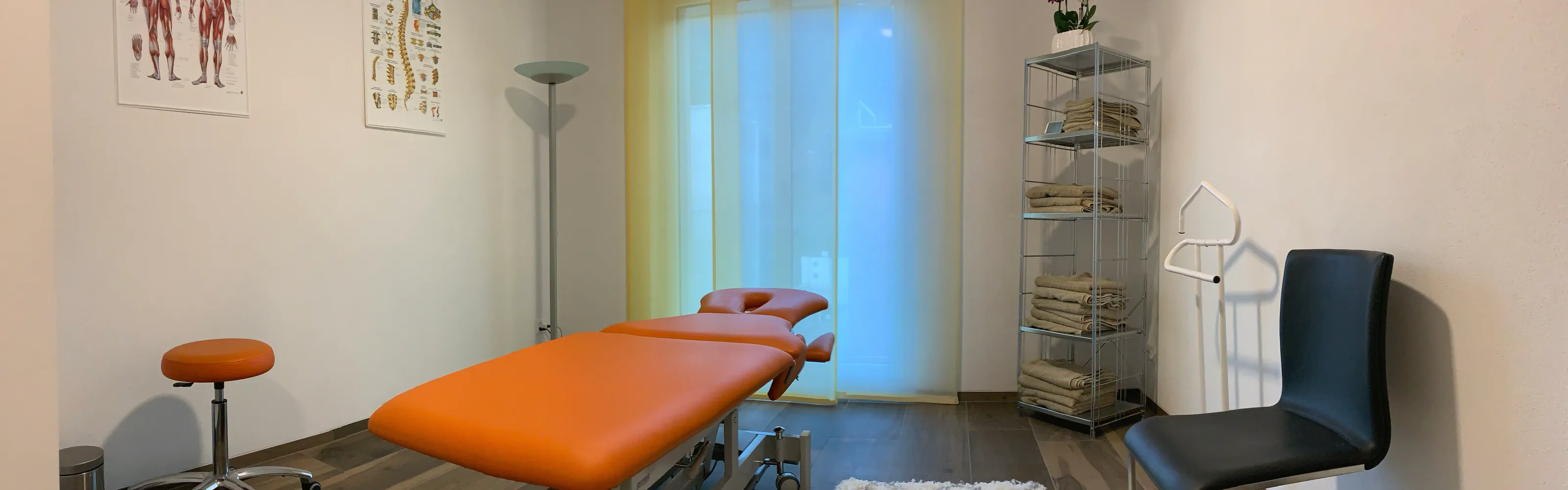 Treatment room with an orange massage table, stool, shelf of towels and two anatomy charts.