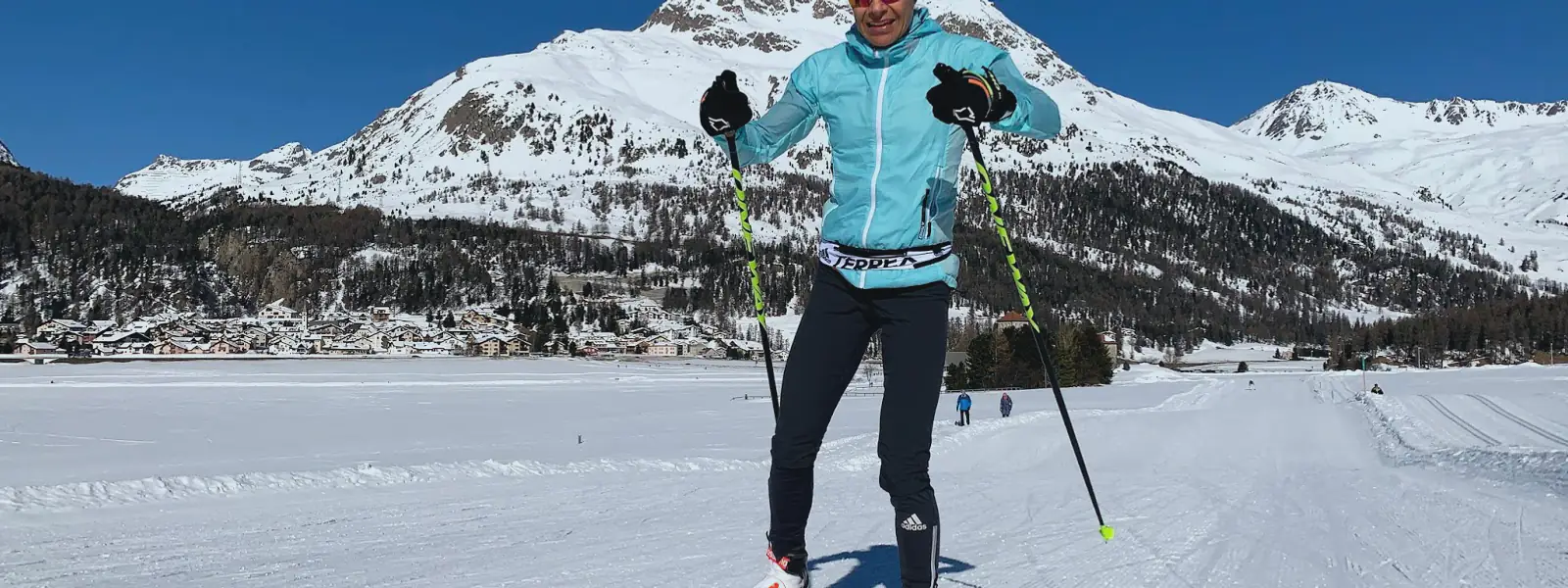A cross-country skier skating on a groomed track below snow-covered mountains and a village.